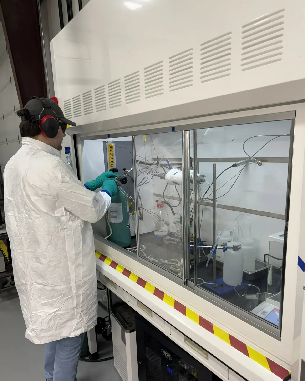 Scientist in a white coat and ear protection working through the raised sash of a fume hood