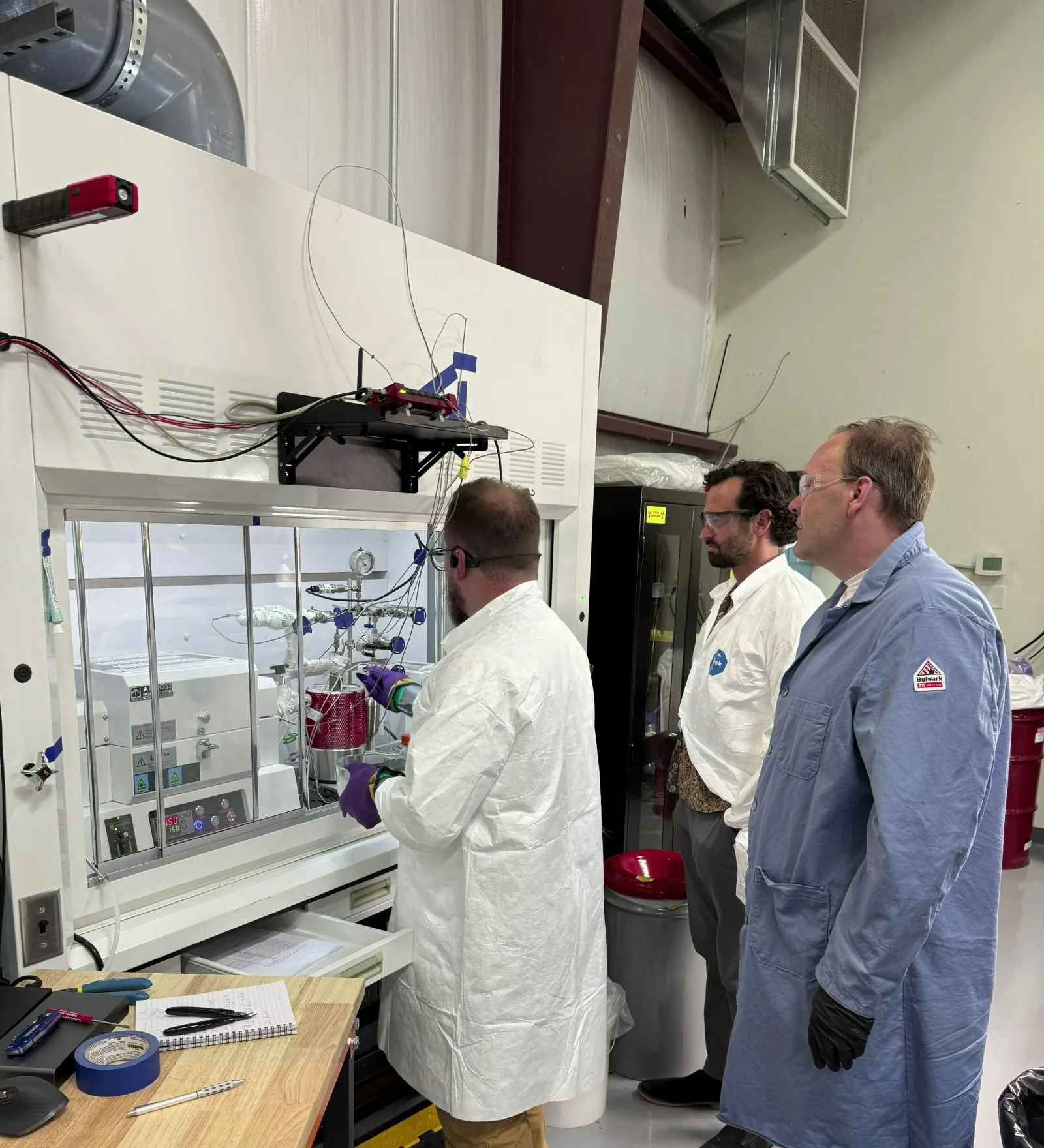 Scientist working at a fume hood while two people in lab coats and safety glasses watch; the exhaust duct rises from the hood to the ceiling