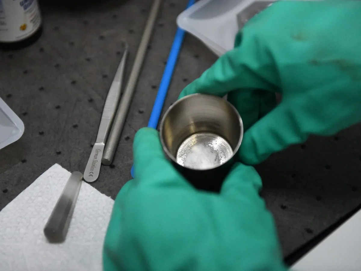 Green-gloved hands holding a small steel crucible over a tray, spatula beside it