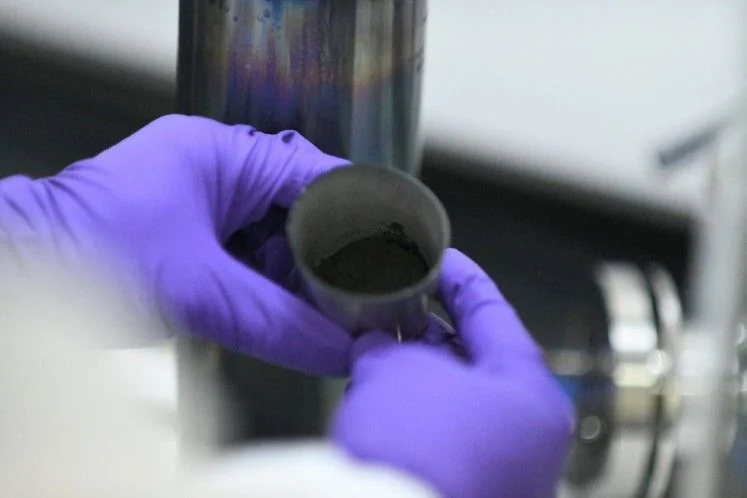 Gloved hand holding a small cup of black uranium dioxide powder