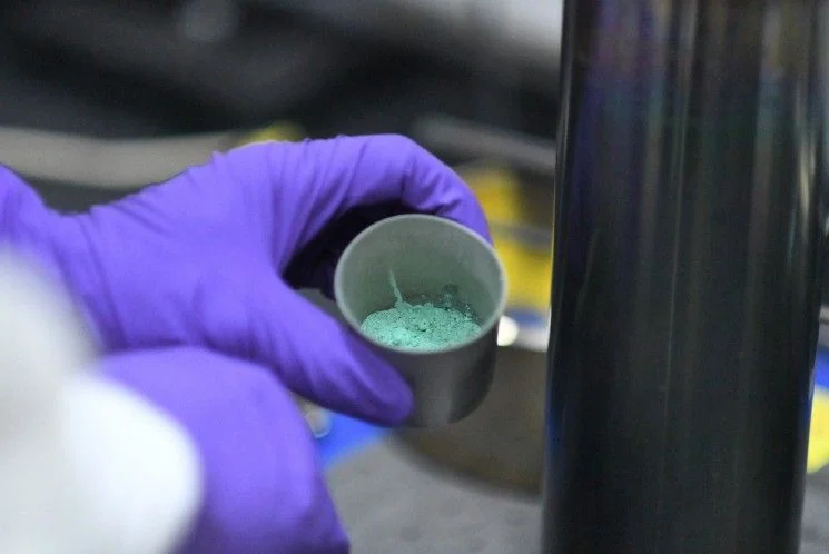 Gloved hand holding a small cup of pale green uranium tetrafluoride powder