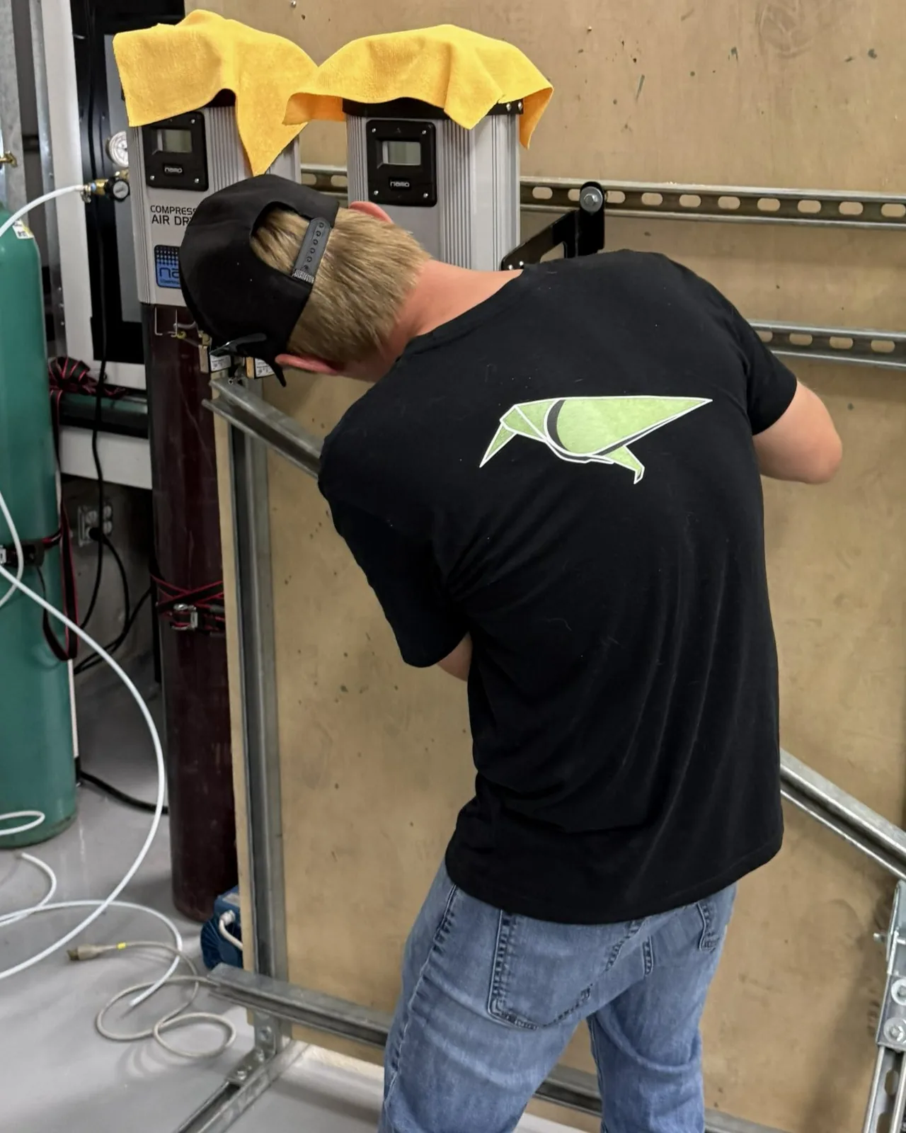 A team member in a black Raven-Flint shirt building the gas-drying rig, plumbing compressed-air dryers onto a strut wall beside gas cylinders
