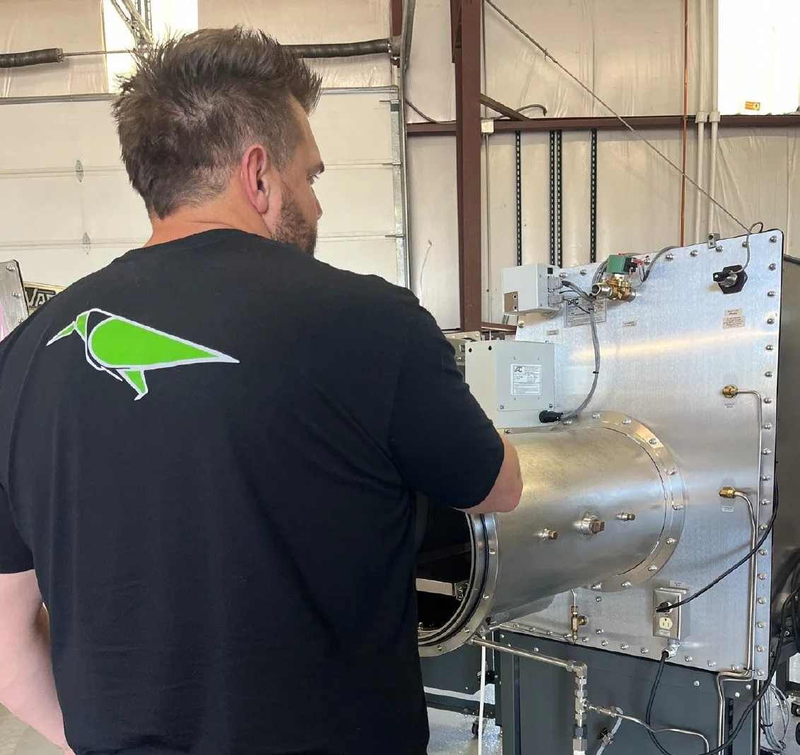 A team member in a black Raven-Flint shirt, seen from behind, working at the glovebox antechamber