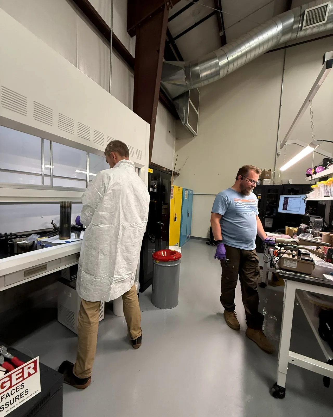 Two scientists at a bench in the laboratory, one in a white coat at the hood, one at a laptop
