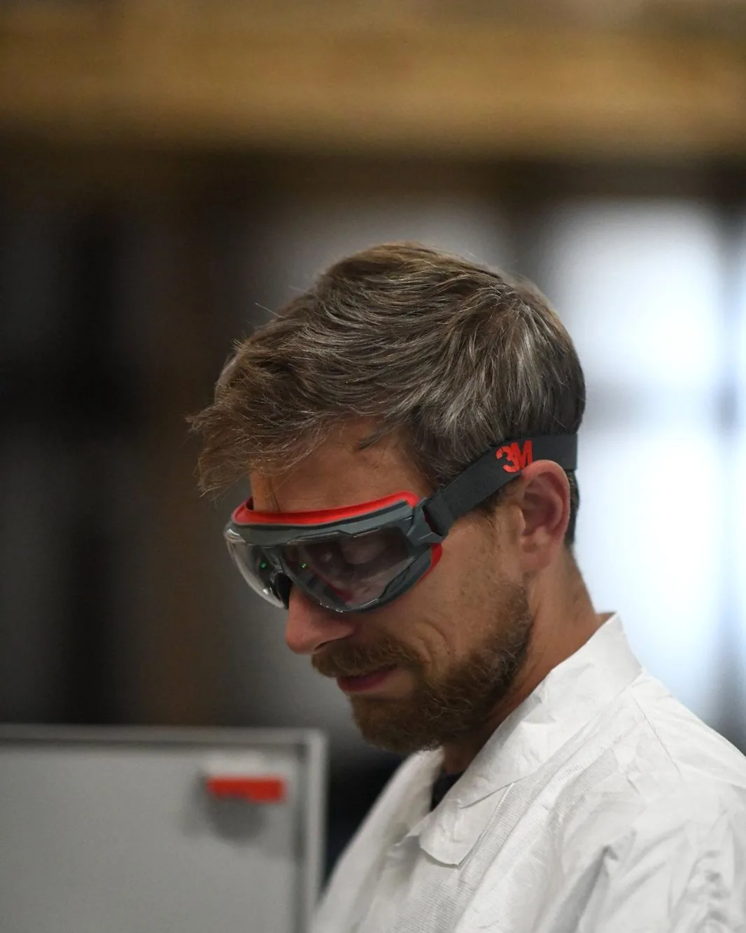 A scientist in a white coat and goggles, in profile, at an instrument