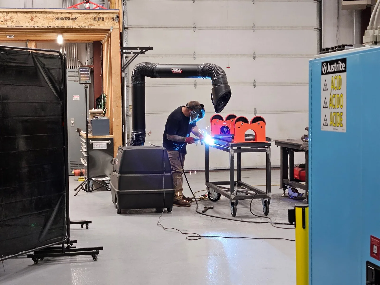 A welder at a steel fixture under a fume-extraction arm, welding screens behind, a blue acid cabinet in the foreground