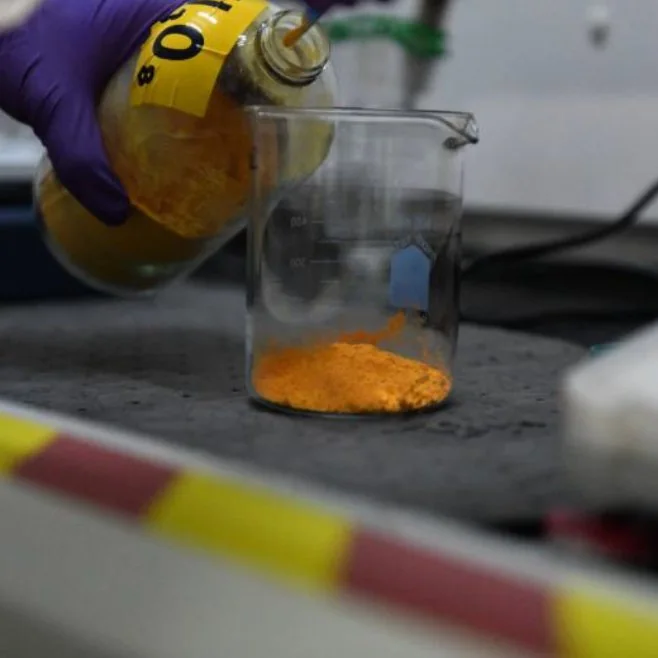 Gloved hand pouring orange-yellow uranium ore concentrate from a labeled jar into a glass beaker